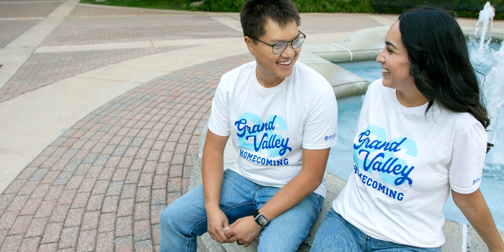 A couple smiles wearing the GVSU Homecoming T-shirts on campus sitting beside a fountain